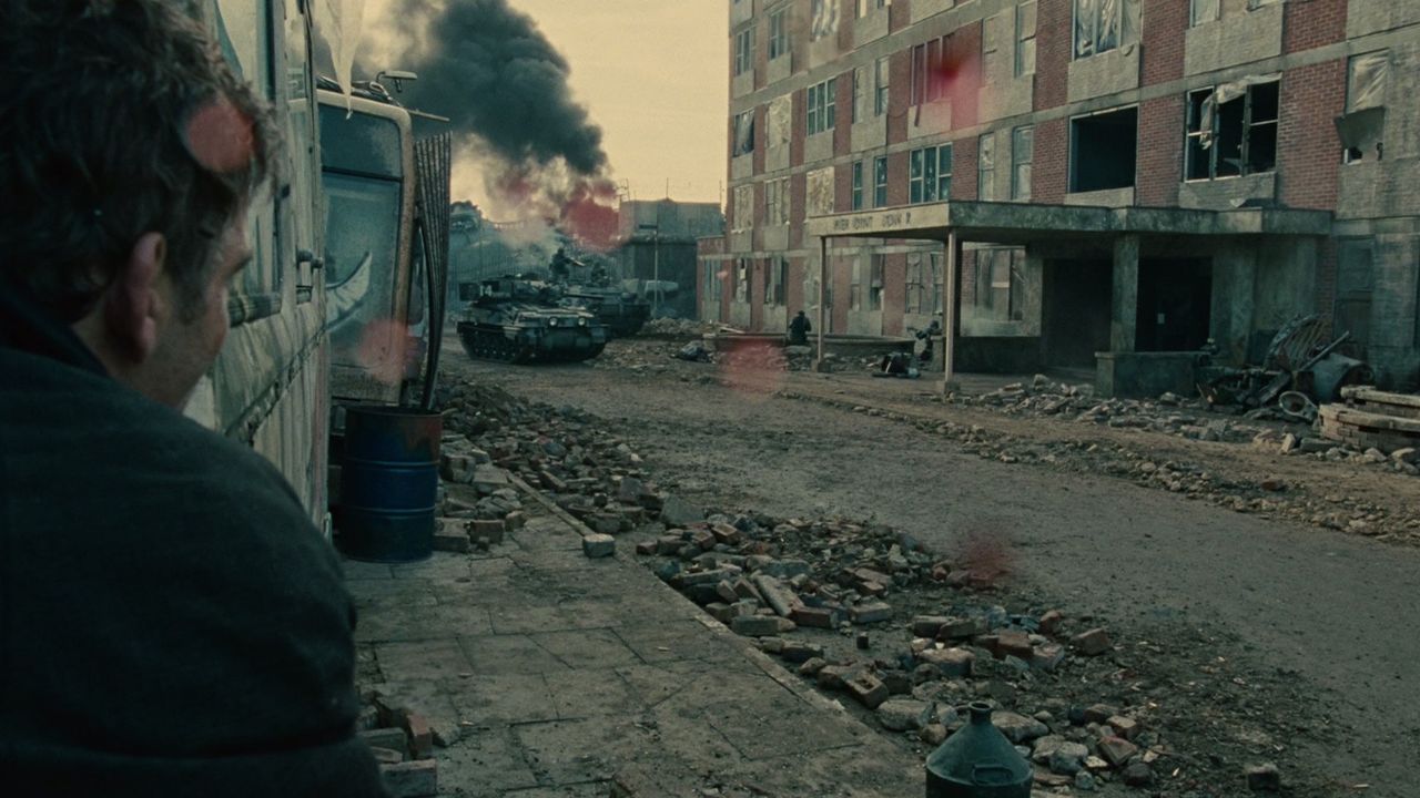 Auf dem Bild ist eine zerstörte Stadt zu sehen. In der Mitte der Straße sind Panzer und eine Rauchwolke zu sehen. Links im Bild kauert ein Mann an der Wand und schaut zu den Panzern rüber