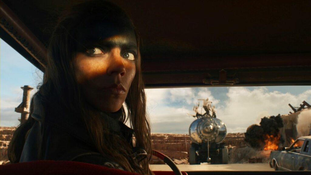 Furiosa: A Mad Max Saga: Auf dem Bild sieht man eine Frau, die am Steuer eines Autos sitzt und sich gerade nach hinten umwendet. Vor ihr ist eine Art Mauer, gegen die ein Truck gefahren ist, auf dem bleiche Menschen stehen. Eine Top-Fahrzeug-Actionszene.