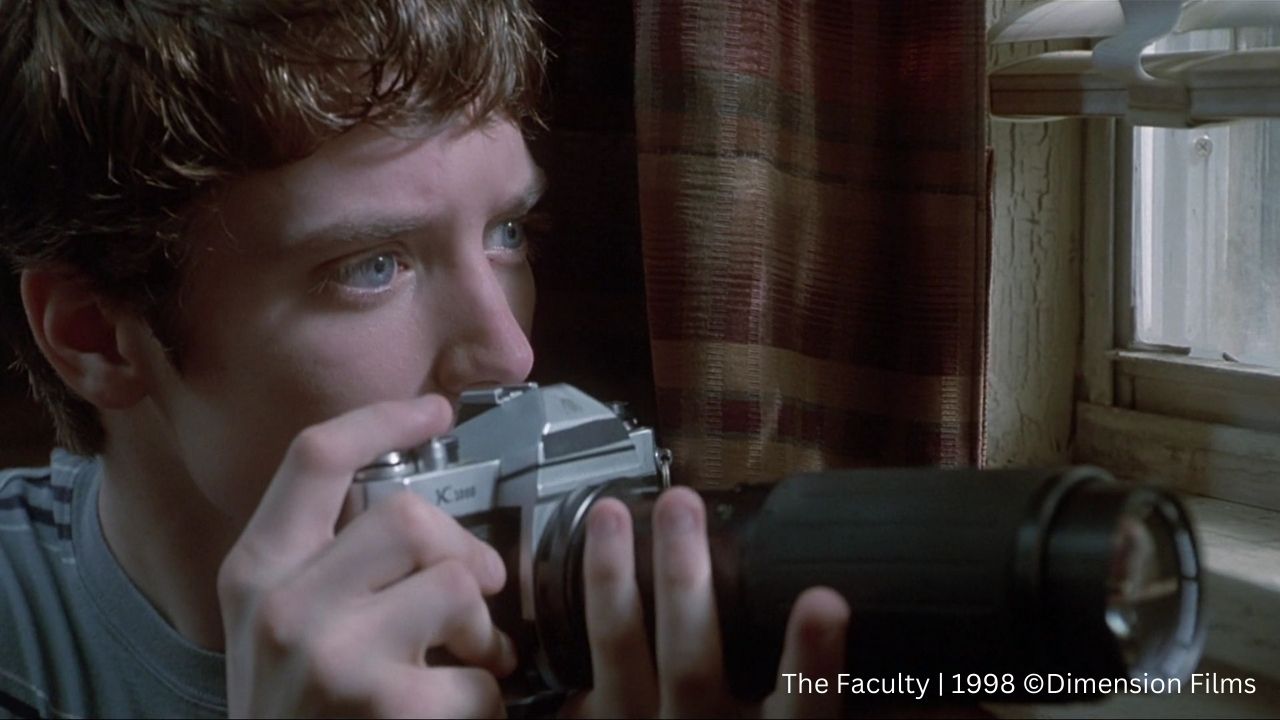 Kinderdarsteller: Auf dem Bild hat ein junger Mann eine Kamera in der Hand und versteckt sich hinter einem Vorhang. Sein Blick geht nach draußen aus einem Fenster
