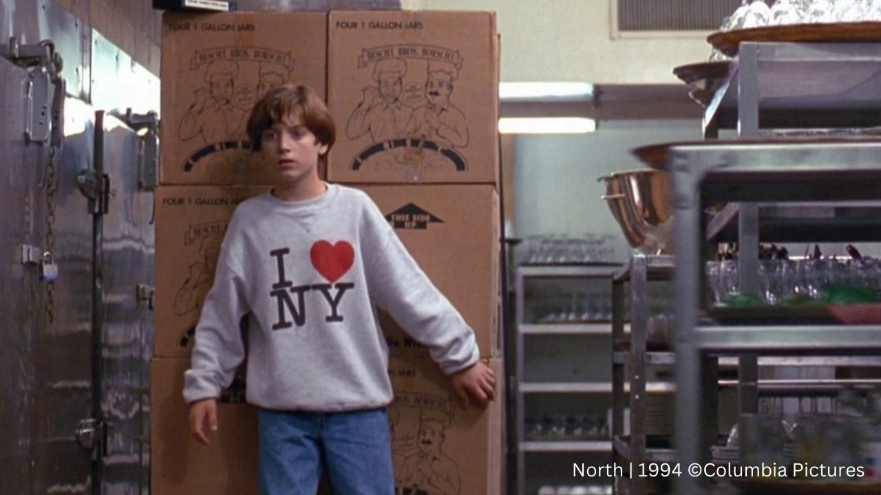 Auf dem Bild ist ein Junge zu sehen, der einen "I Heart NY" Pullover trägt. Er steht in einem Lagerraum vor Pappkisten