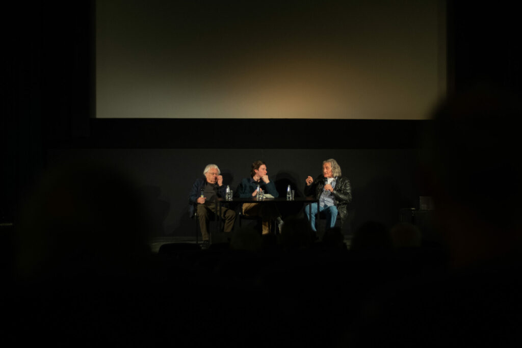 Sehsüchte Filmfestival: Das Bild zeigt drei Menschen vor einer Kinoleinwand, die am Tisch sitzen und diskutieren