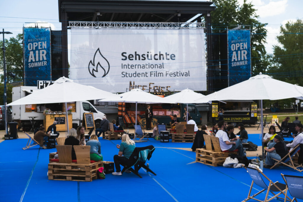 Das Bild zeigt eine große Leinwand mit dem Schriftzug Sehsüchte International Student Film Festival. Davor sind weiße Schirme zu sehen unter denen Menschen in Gruppen auf Palettensesseln sitzen