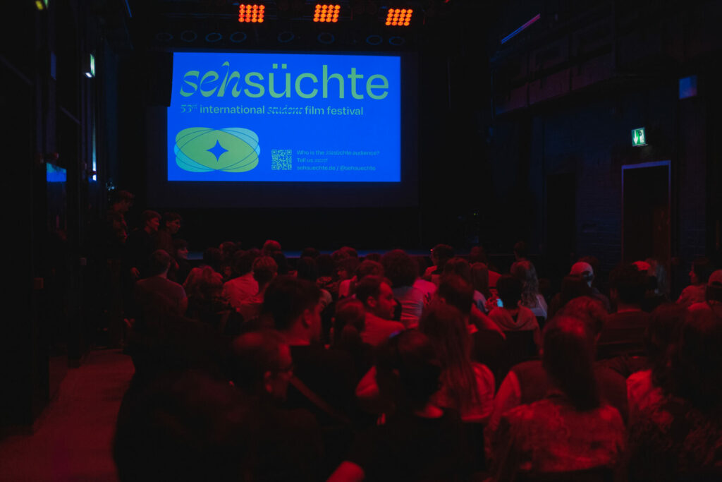 Sehsüchte Filmfestival: Auf dem Bild sieht man einen Kinosaal. Menschen sitzen im roten Licht vor einer Leinwand, die das Gelb Blaue Logo des Sehsüchte Filmfestivals zeigt