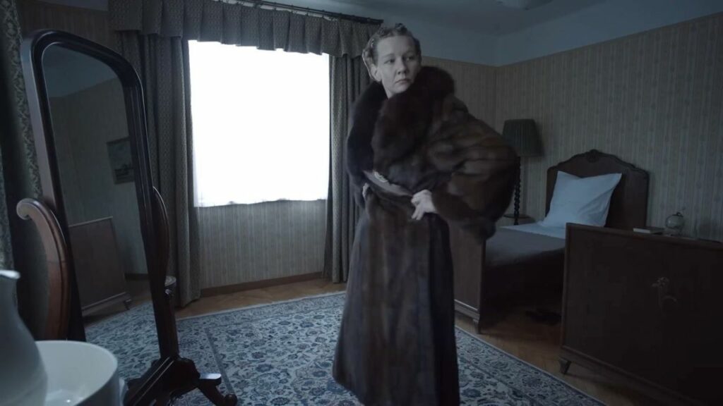 Auf dem Bild ist eine Frau zu sehen, die in einem Schlafzimmer vor einem Spiegel steht. Sie trägt einen Pelzmantel und beobachtet, wie gut er ihr steht