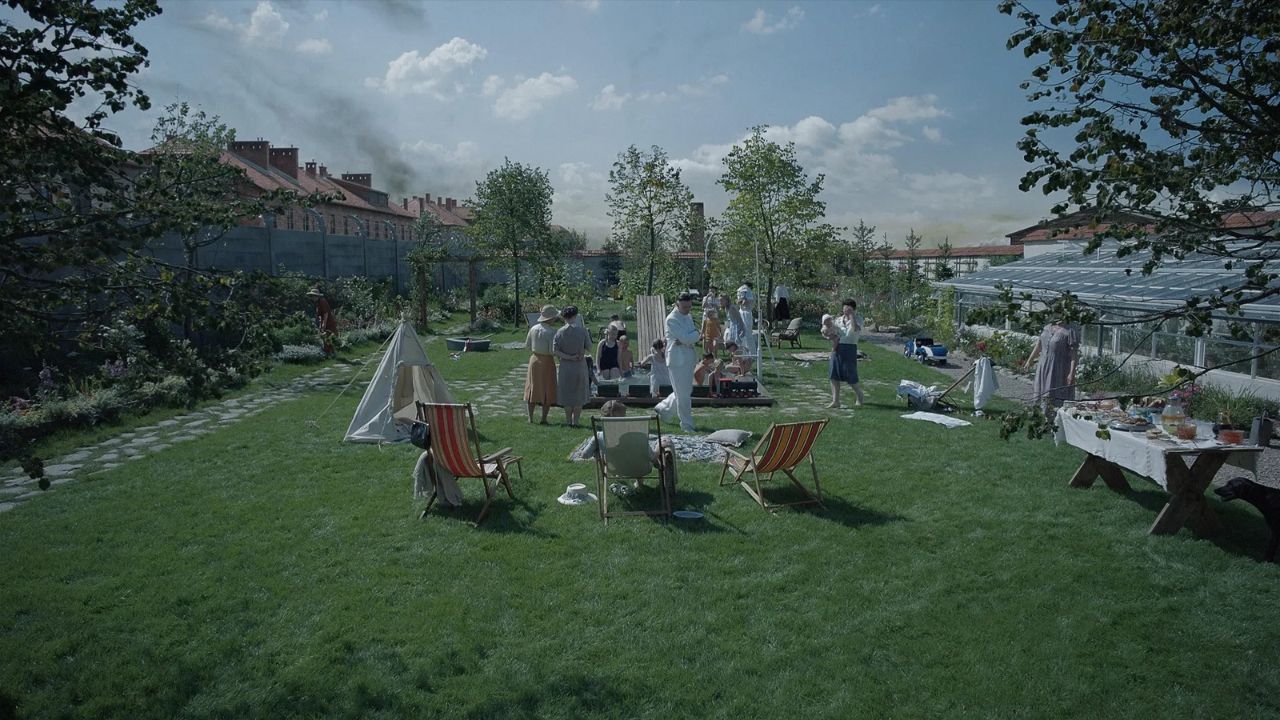 Auf dem Bild ist ein riesiger Garten zu sehen, in der eine Familie ein Fest feiert. Links sieht man rauchende Schornsteine von einer Häuserreihe, die sich hinter einer Mauer befindet