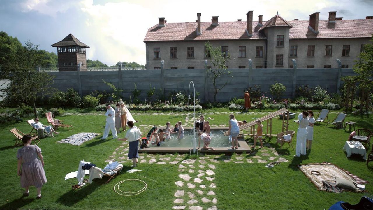 The Zone of Interest: Das Bild zeigt einen schönen Garten, der in der Mitte einen kleinen Pool hat. Darin toben ein paar Kinder. Rundherum stehen Erwachsene. An dem Garten grenzt eine Mauer, die oben Stacheldraht hat. Dahinter ist ein großes Gebäude zu sehen