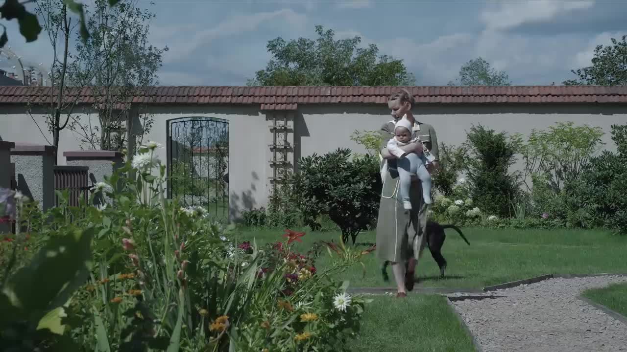 Das Bild zeigt eine Frau, die ein Baby auf dem Arm hält. Sie steht inmitten eines schönen Gartens. Im Hintergrund sieht man eine Mauer mit einem Tor