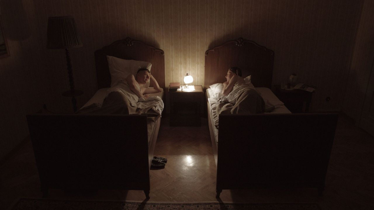 The Zone of Interest: Auf dem Bild ist ein Mann und eine Frau zu sehen, die jeweils in einem Bett liegen. Sie haben jeweils den Arm aufgestützt und schauen sich an. In der Mitte steht ein Nachttisch, auf dem eine Lampe leuchtet