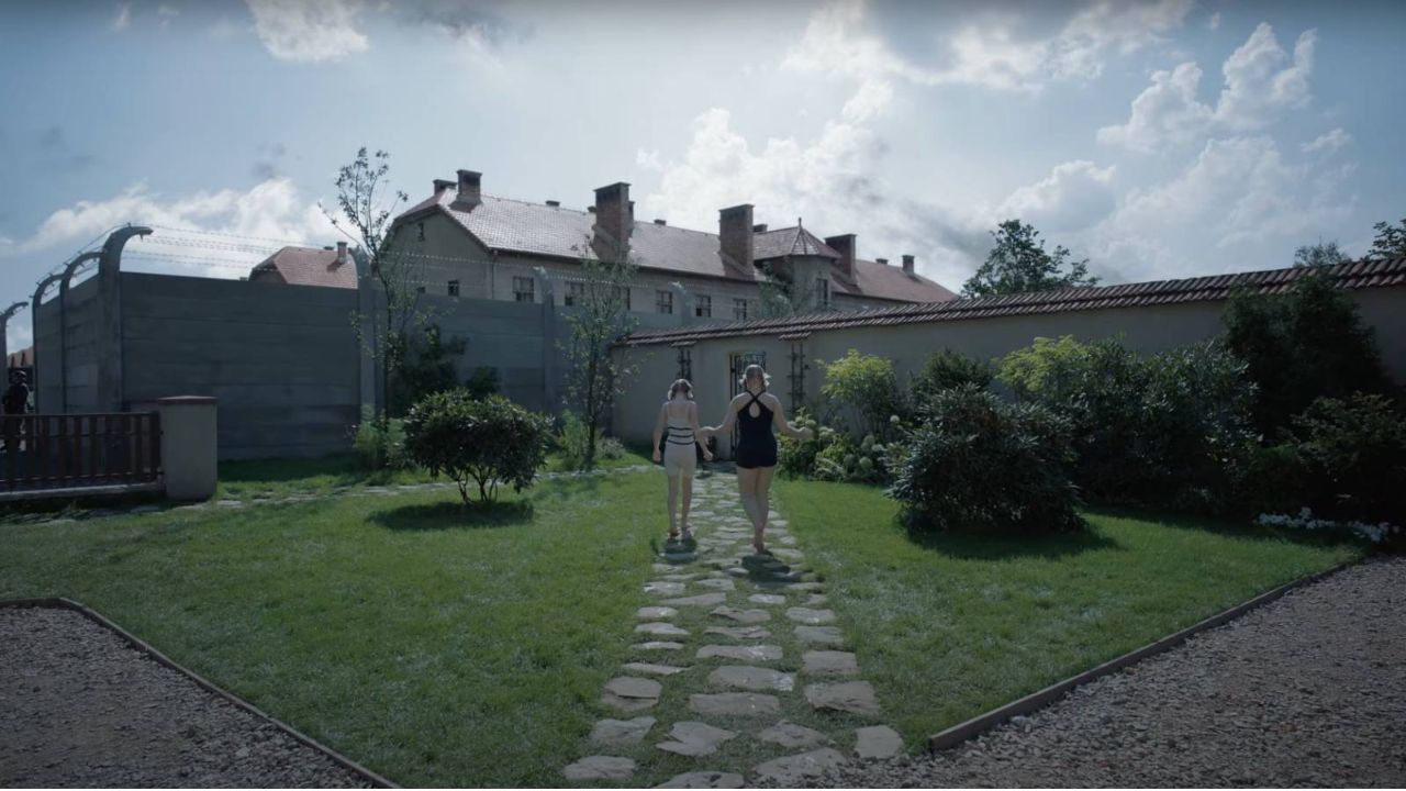 Das Bild zeigt zwei Mädchen im Badeanzug, die einen Weg durch einen Garten gehen. Dahinter ist ein großes Haus zu sehen, das von einer großen Mauer verborgen wird
