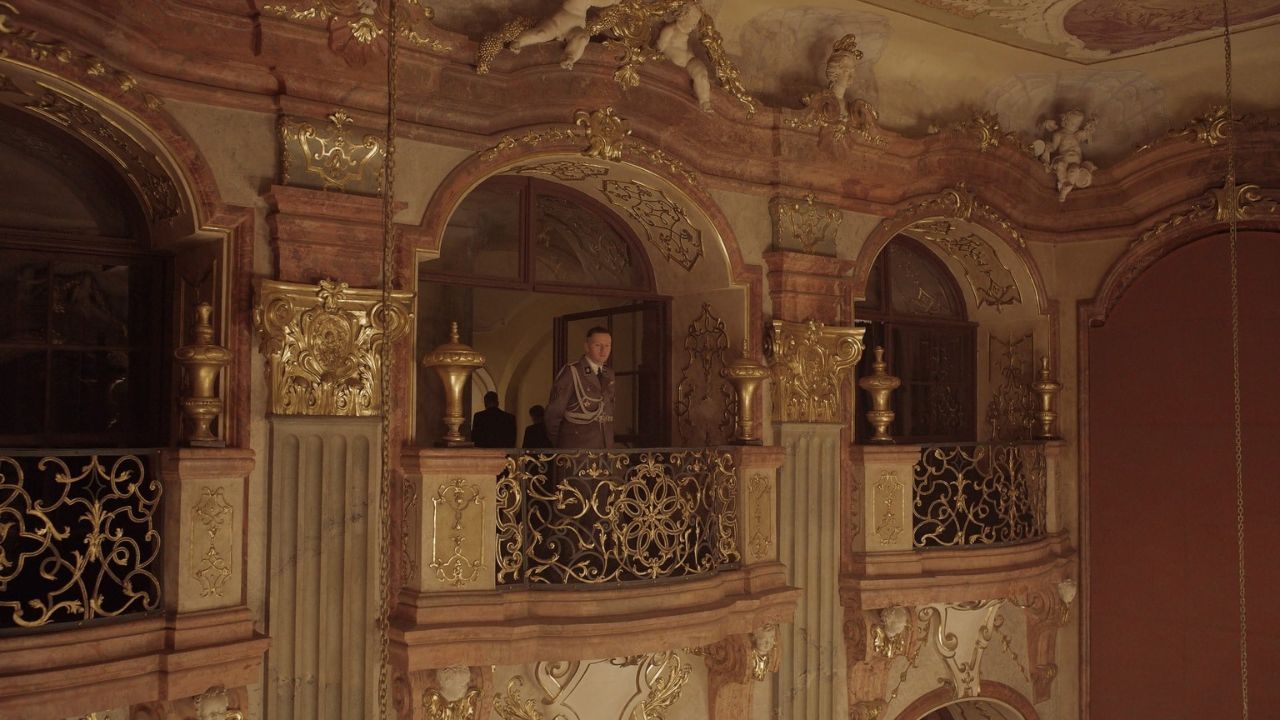 Auf dem Bild ist der Balkon eines prunkvollen Saals zu sehen. An der Brüstung steht ein Mann in Uniform