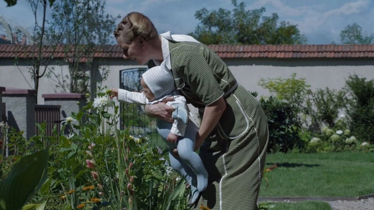 The Zone of Interest: Auf dem Bild ist eine Frau, die ein Baby auf dem Arm hält. Sie steht in einem schönen Garten und lässt das Baby eine Blume zupfen