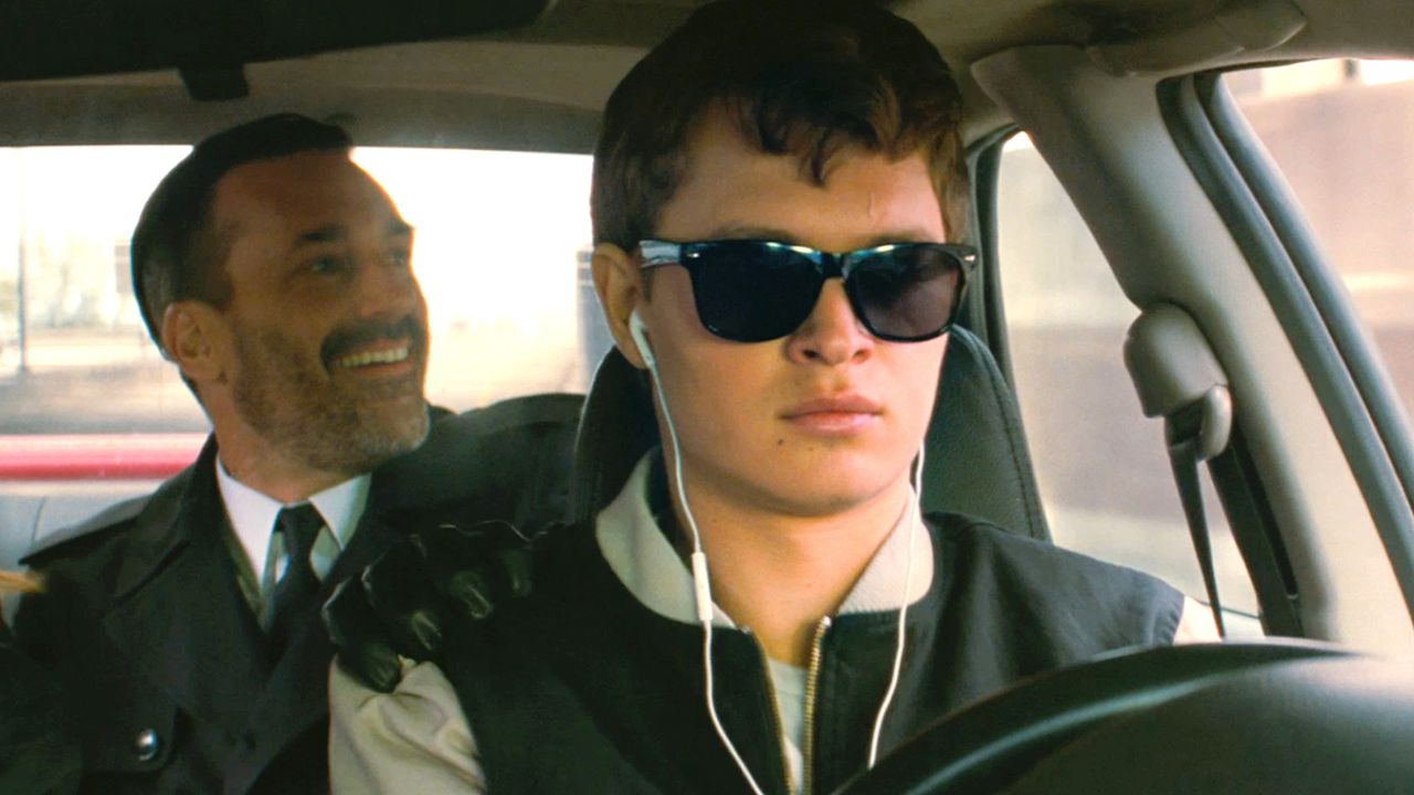 Auf dem Bild ist ein junger Mann am Steuer eines Autos zu sehen. Er trägt Kopfhörer und eine Sonnenbrille. Hinten im Auto sitzt ein Mann im Anzug, der freudig lacht