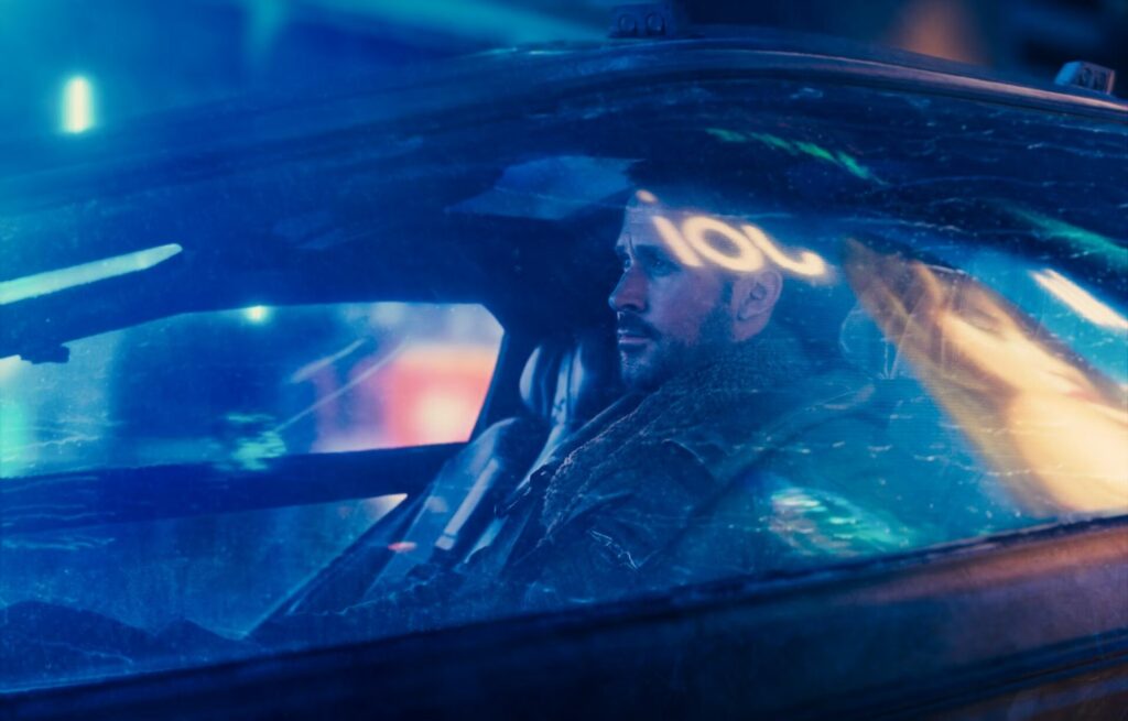 Blade Runner 2049: Im Neon Licht! Auf den Bild sitzt ein Mann im Auto. Er ist vom blauen Neonlicht umgeben. Man sieht den Mann von außen gefilmt auf der Scheibe ist ein Schriftzug mit der Aufschrift "Joi" spiegelverkehrt zu sehen