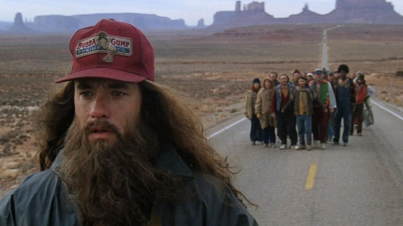 Forrest Gump: Das Bild zeigt vorne links einen Mann mit langem Bart und langen Haaren. Er trägt ein rotes Cap mit der Aufschrift Bubba Gum Shrimp & Co. Hinter ihm auf der Straße steht eine Gruppe Menschen. Im Hintergrund sind Berge zu sehen