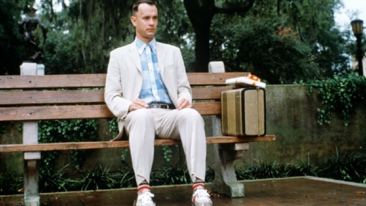 Auf dem Bild ist ein Mann zu sehen, der auf einer Parkbank sitzt. Neben ihm steht ein Koffer, darauf eine Schachtel Pralinen