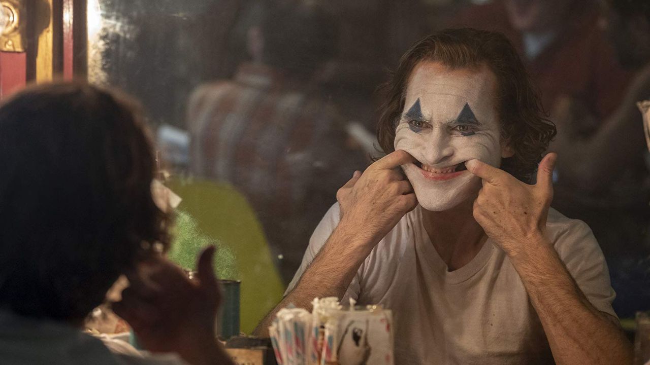 Joker: Auf dem Bild ist ein Mann zu sehen, dessen Gesicht teilweise als Clown geschminkt ist. Er schaut in der Garderobe gerade in den Spiegel und zieht sich mit den Fingern ein lächelndes Gesicht