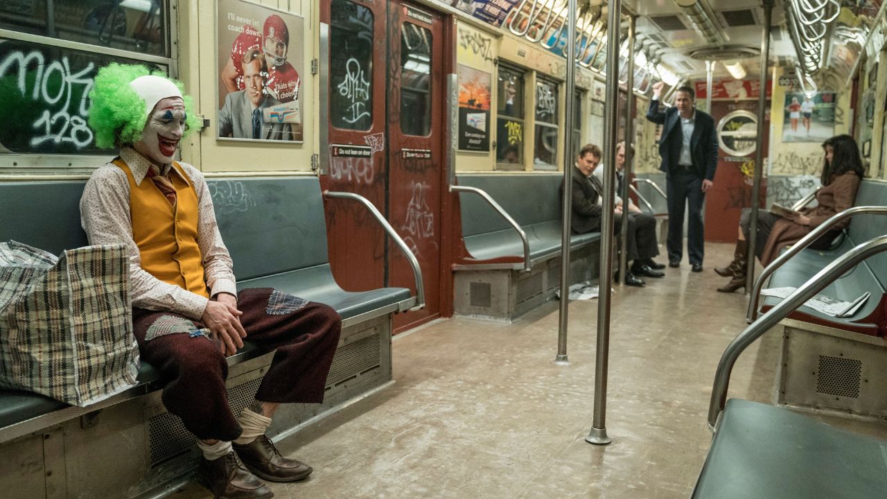 Das Bild zeigt ein U-Bahn Abteil. Auf einer Bank am Fenster sitzt ein Clown, der vor sich hin lacht. Im hinteren Teil sind Menschen zu sehen, die zu dem Clown hinüber blicken