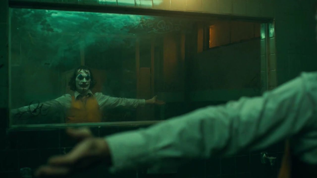 Joker: Auf dem Bild ist ein Mann mit einem Clown Gesicht, der vor dem Spiegel steht. Man sieht nur sein Spiegelbild im Spiegel. Er trägt ein weißes Hemd, eine gelbe Weste und breitet die Arme aus
