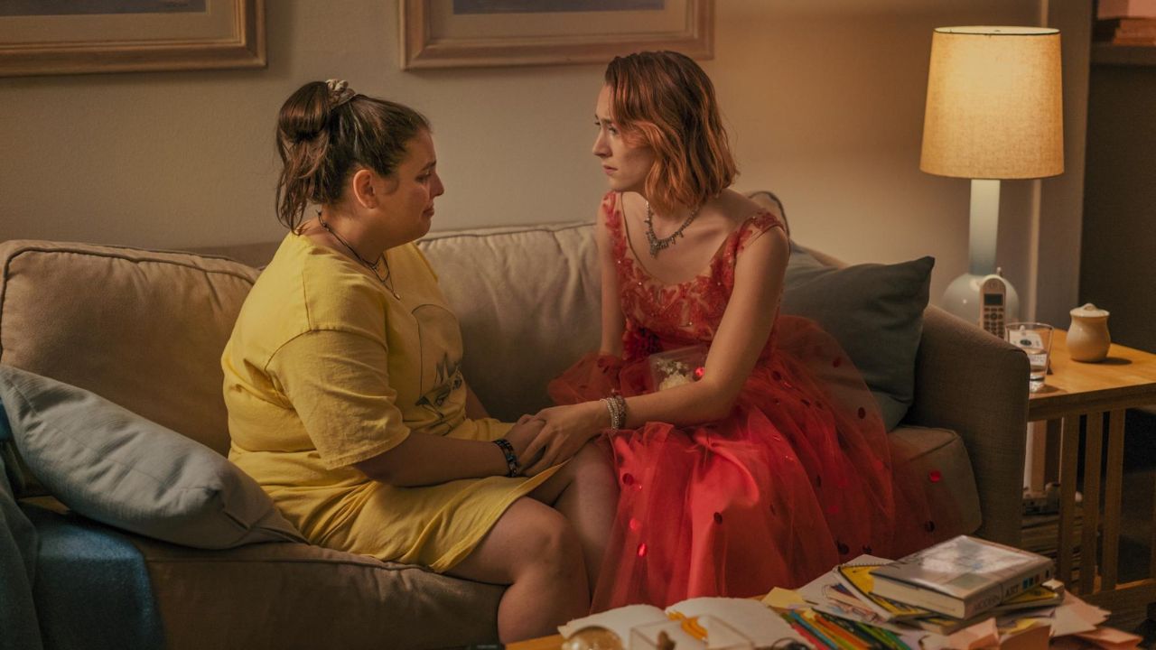 Lady Bird: Das Bild zeigt zwei junge Frauen, die auf einer Couch sitzen. Die eine trägt ein rotes Ballkleid, die andere ein gelbes Longshirt. Die Person im Longshirt weint gerade, die andere tröstet sie