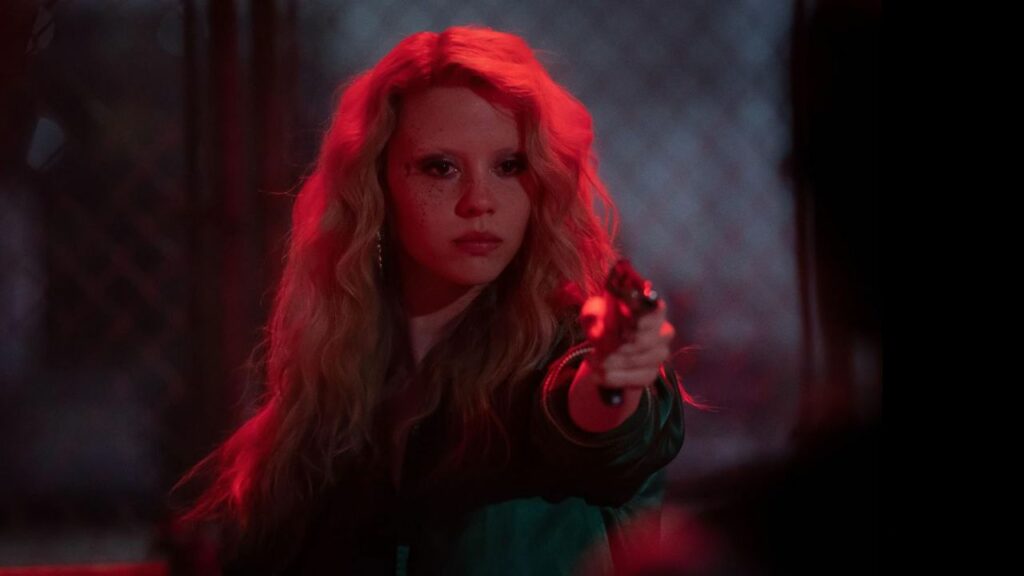 MaXXXine: Das Bild zeigt eine junge Frau, die im roten Lichtschein steht und die Waffe auf eine Person vor ihr richtet