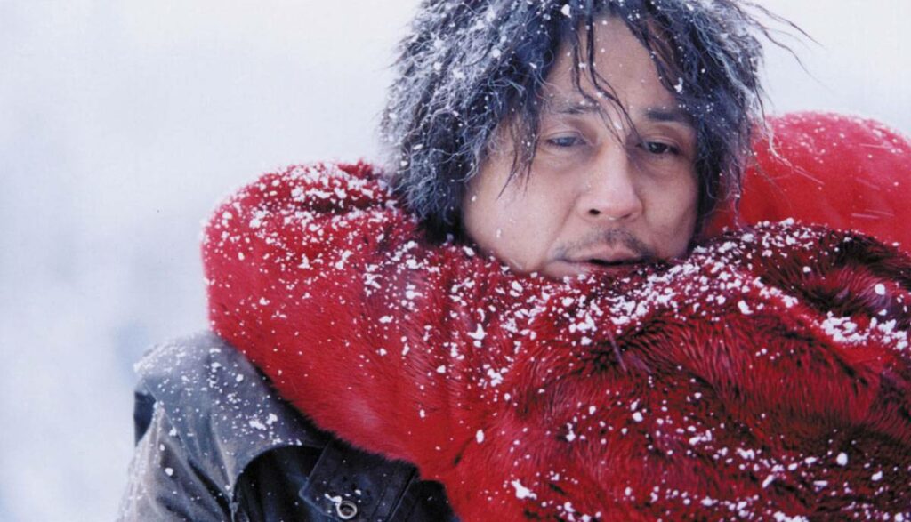 Plot-Twist: Auf dem Bild ist ein Mann zu sehen, der eine Person in einem roten Mantel umarmt. Er blickt traurig drein, sein Haar ist mit Schnee bedeckt