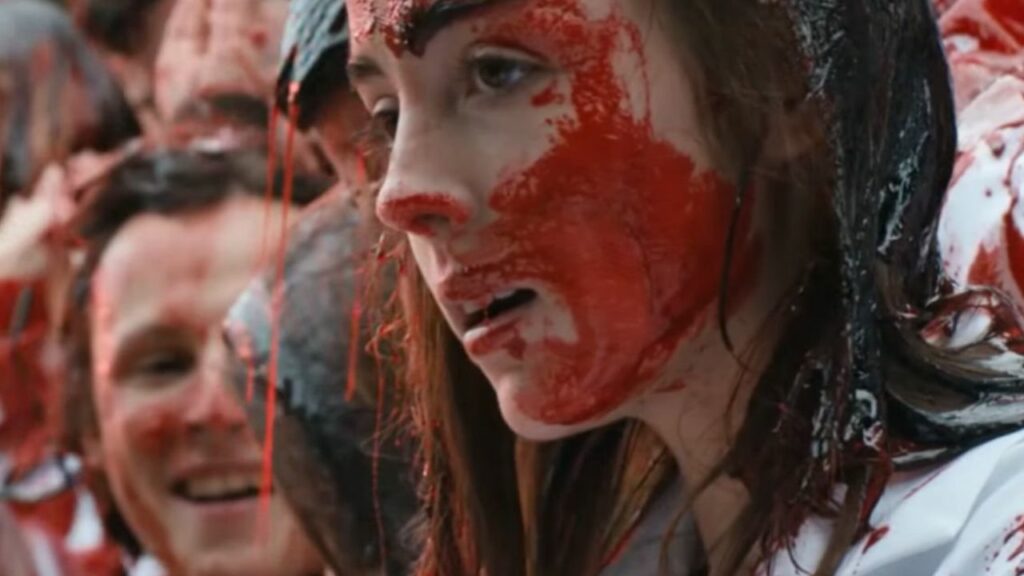Auf dem Bild ist eine Gruppe junger Menschen, die mit roter Farbe bedeckt sind. Im Profil im Vordergrund ist eine junge Frau mit viel roter Farbe im Gesicht