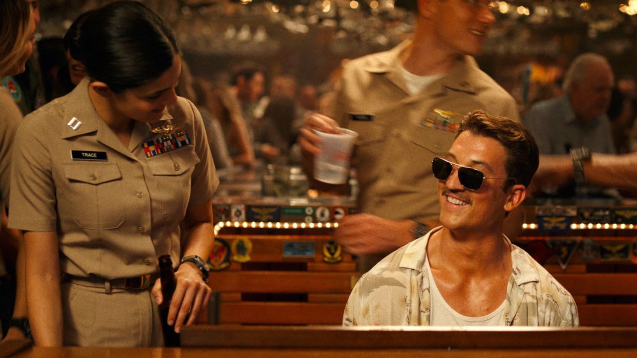 Top Gun: Maverick: Das Bild zeigt einen jungen Mann mit Fliegerbrille am Klavier. Neben ihm steht eine Frau in Uniform. Hinter ihm geht ein Mann in Uniform und Brille. Er trägt einen Becher mit Bier darin