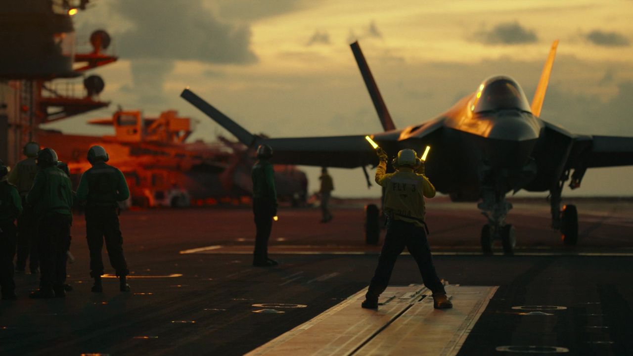 Top Gun: Maverick: Auf dem Bild ist eine Flugbahn zu sehen auf der ein Flugzeug steht. Ein Mann steht mit Leuchten davor. Neben ihm ist eine weitere Personengruppe. Im Hintergrund sieht man einen Kran