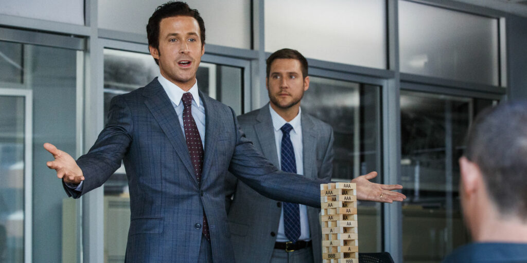 Die 5 besten Filme, die auf wahren Begebenheiten basieren: Auf dem Bild ist ein Mann im Anzug zu sehen, der vor einer Person sitzt und ihr etwas mit ausgebreiteten Armen erklärt. Vor ihm auf den Tisch steht ein Jenga Turm mit vielen As und Bs beschriftet. Neben ihm steht noch ein Mann im Anzug, der ihm aufmerksam zuhört. Dahinter ist eine Glastürenfront zu sehen