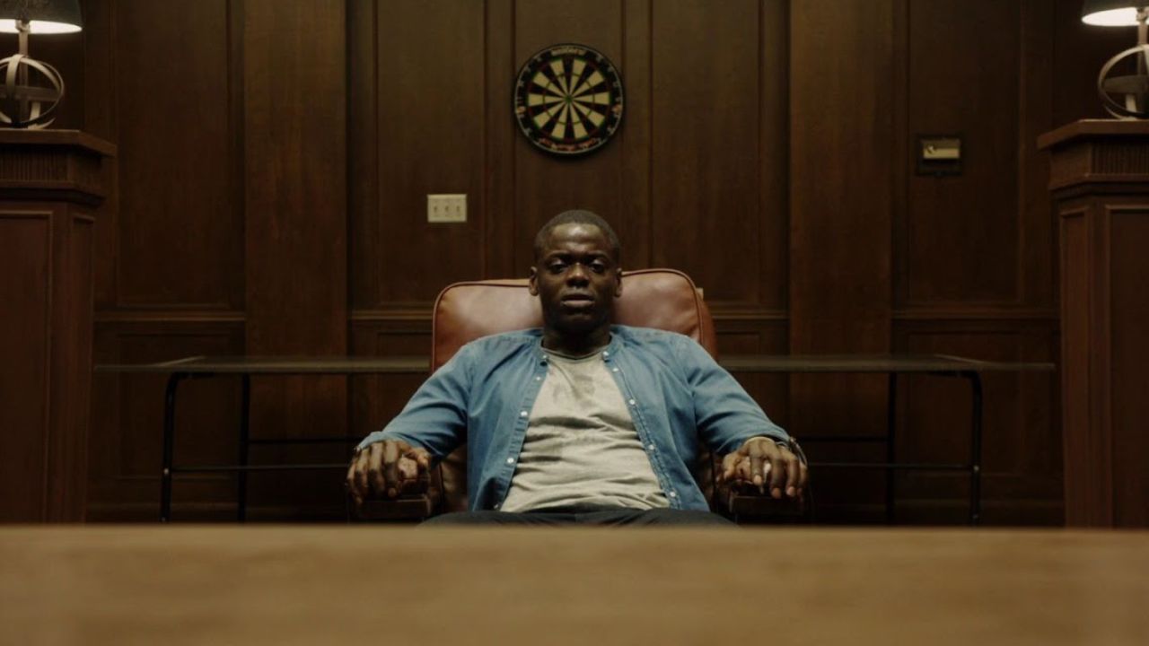 Top Lieblings-Horrorfilme: Get Out: Auf dem Bild sitzt ein Mann in einem Sessel an einem Tisch. Hinter ihm ist eine Holzwand mit einer Dartscheibe | Top 5 Moderne Horrorfilme