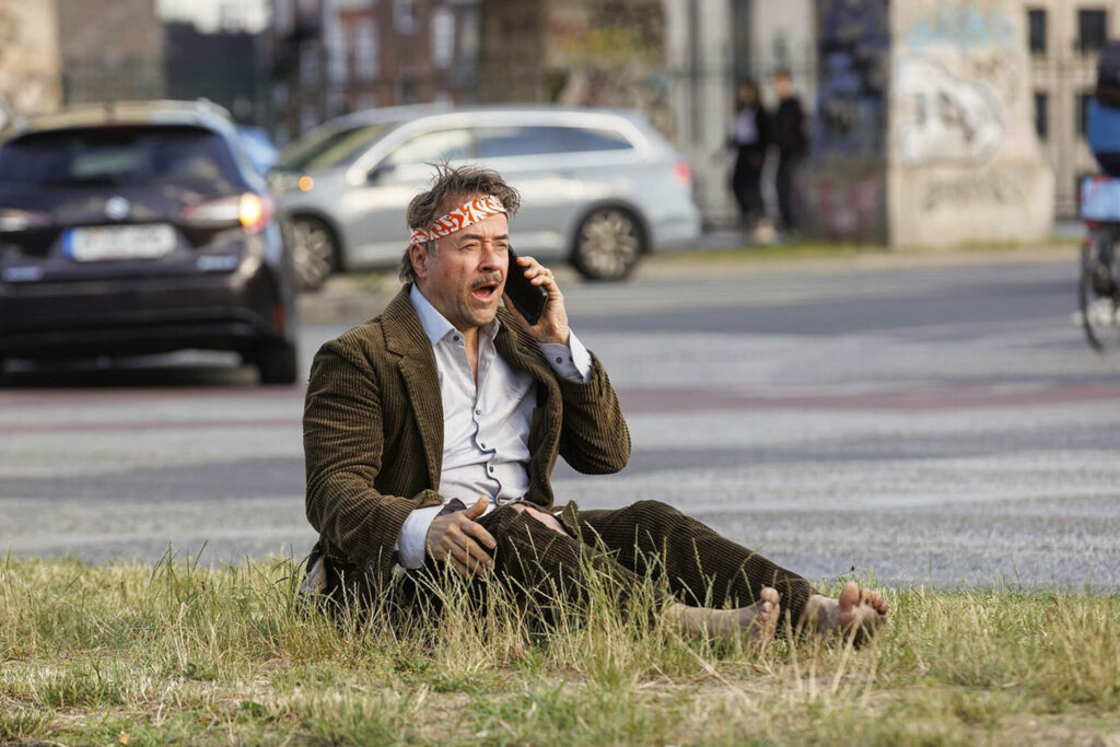 Das Bild zeigt einen Mann, der ohne Schuhe auf einer Grünfläche an der Straße sitzt und mit dem Handy telefoniert