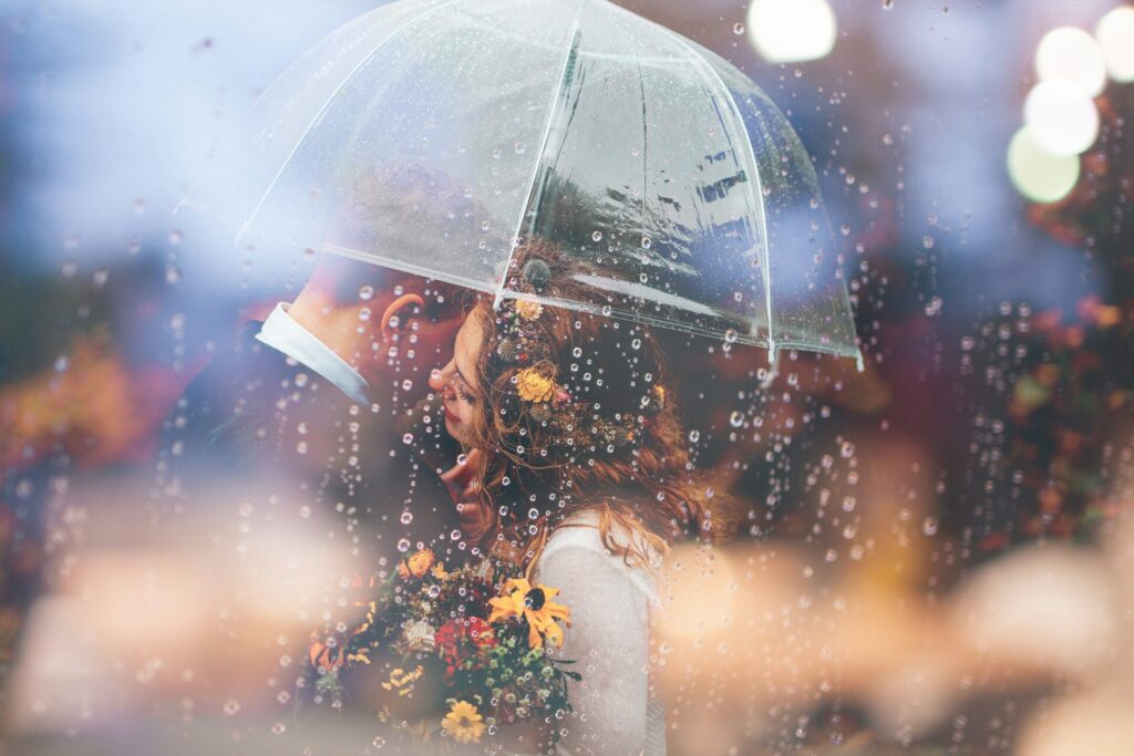 Ein verliebtes Paar steht unter einem transparenten Regenschirm, geschützt vor sanft fallendem Regen. Die Frau hält einen bunten Blumenstrauß, und im Hintergrund schimmert ein herbstliches Bokeh, das eine romantische und warme Atmosphäre erzeugt. | Die 5 liebsten Buch Charaktere