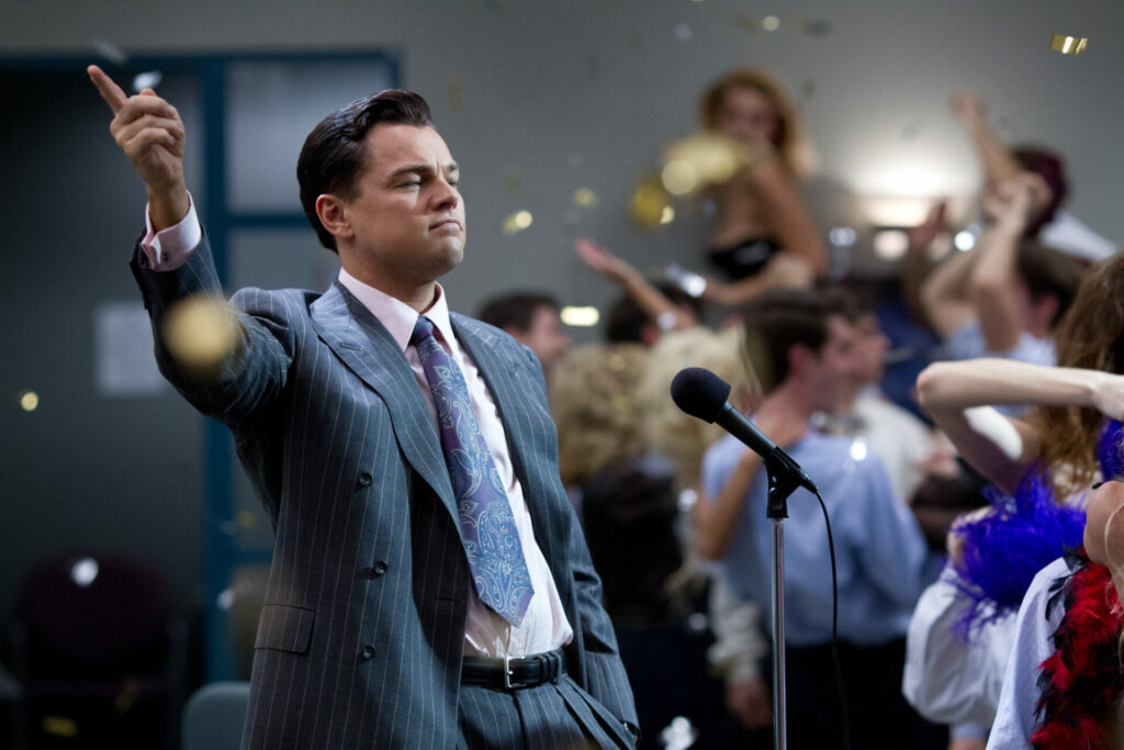 The Wolf of Wall Street: Auf dem Bild ist ein Mann im Anzug, der vor einem Mikrofon steht, die Hand hebt und mit einem Finger zeigt und einen zufriedenen Gesichtsausdruck hat. Im Hintergrund sieht man goldenes Konfetti und tanzende Menschen | Media Monday #700