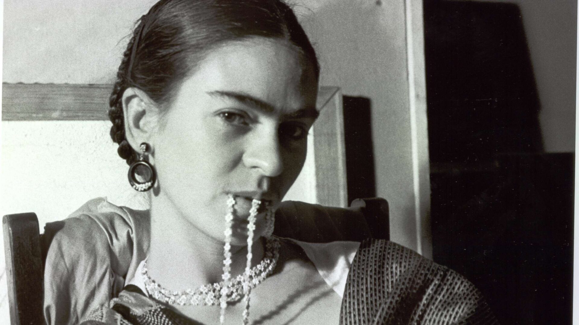 Frida: Das Bild zeigt eine Schwarz-Weiß Aufnahme von Frida Kahlo, sitzend auf einem Stuhl. Kek hat sie ihre Kette in den Mund genommen und blickt herausfordernd in die Kamera