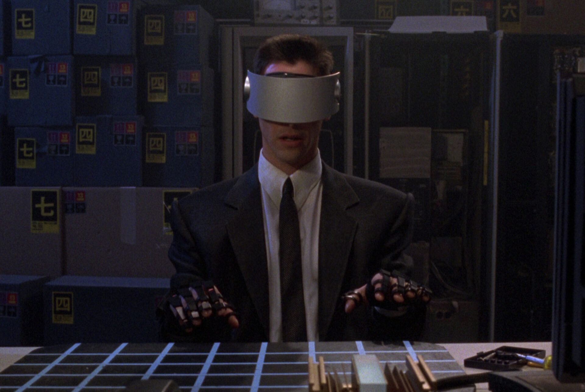 Auf dem Bild ist Johnny Mnemonic, der eine Cyberbrille trägt, dazu Handschuhe und an einem Tisch sitzt