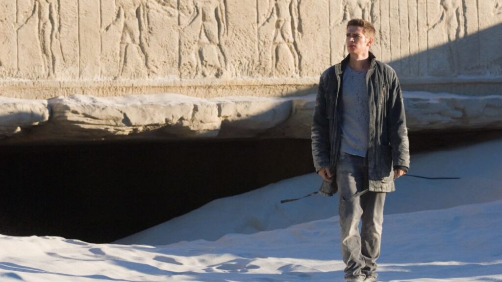 Jumper: Das Bild zeigt einen Mann, der in einer sandigen Wüstenumgebung vor einer alten Steinwand mit ägyptischen Hieroglyphen steht. Er trägt eine dunkle Jacke, ein graues Oberteil und Jeans. Der Ausdruck auf seinem Gesicht wirkt nachdenklich oder beeindruckt und das Licht fällt seitlich auf die Szene, wodurch die Hieroglyphen auf der Wand gut sichtbar hervorgehoben werden. | Top 5 Superkräfte