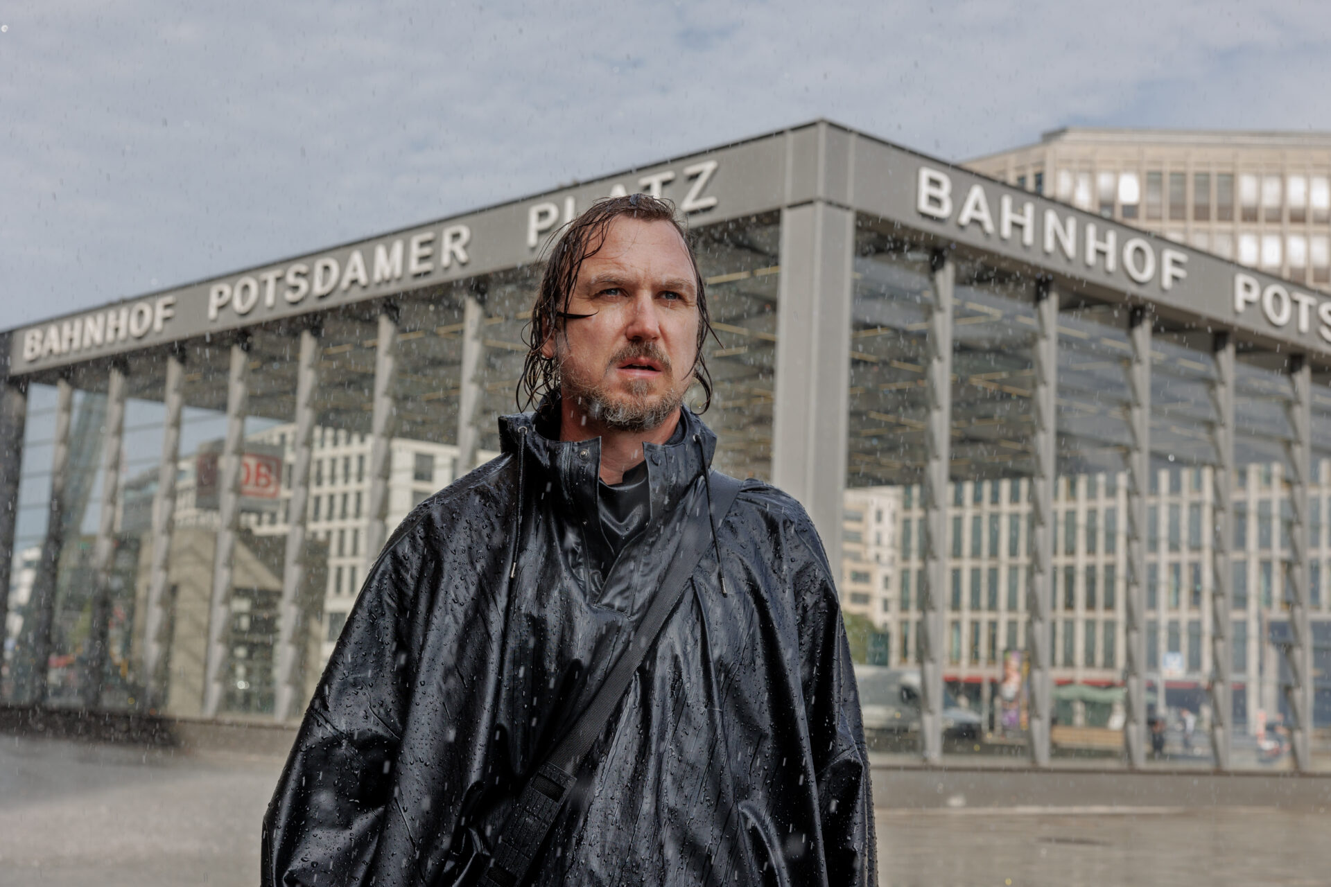 Das Bild zeigt einen Mann vor dem Bahnhofsgebäude des Potsdamer Platzes. Er trägt eine Regenjacke und ist nass geregnet. Es regnet noch immer