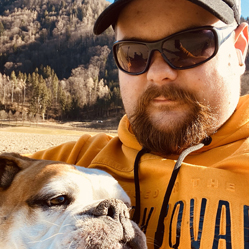 Ein Mann mit Bart, Sonnenbrille und schwarzer Cap hält eine Bulldogge im Arm. Er trägt einen orangefarbenen Hoodie mit schwarzem Schriftzug. Im Hintergrund ist eine hügelige Landschaft mit kahlen und immergrünen Bäumen unter blauem Himmel zu sehen.
