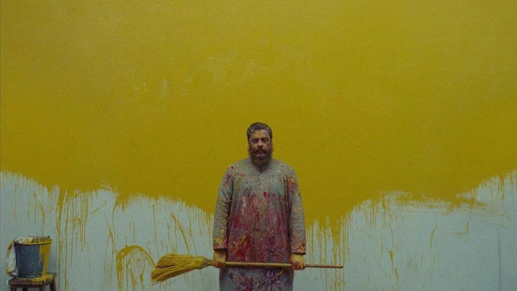 Das Bild zeigt einen Mann im Malerkittel, der voll mit Farbsprenkeln ist. Er steht vor einer halb gelb gestrichenen Wand, hält einen Besen, der mit gelber Farbe voll ist, in den Händen und nebendran steht auf einem Hocker ein Eimer mit gelber Farbe