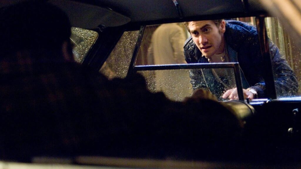 Das Bild zeigt einen Mann, der zu jemand anderen durch ein verregnetes, geöffnetes Autofenster spricht