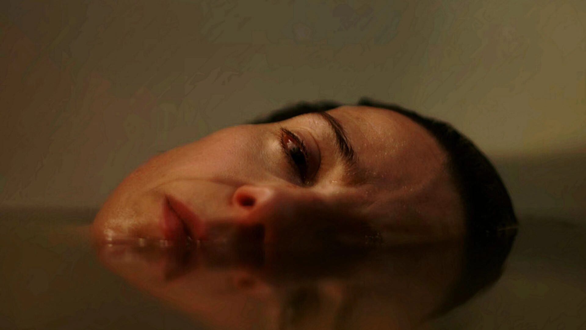 Nahaufnahme eines Gesichts, das teilweise seitlich in Wasser eingetaucht ist. Ein Auge, die Nase und der Mund sind knapp über der Wasseroberfläche sichtbar. Die Wasseroberfläche spiegelt den untergetauchten Teil des Gesichts. Das Licht ist gedämpft und wirft sanfte Schatten und Highlights auf die Haut. Die Szene wirkt ruhig, aber intensiv und vermittelt eine emotionale Spannung.