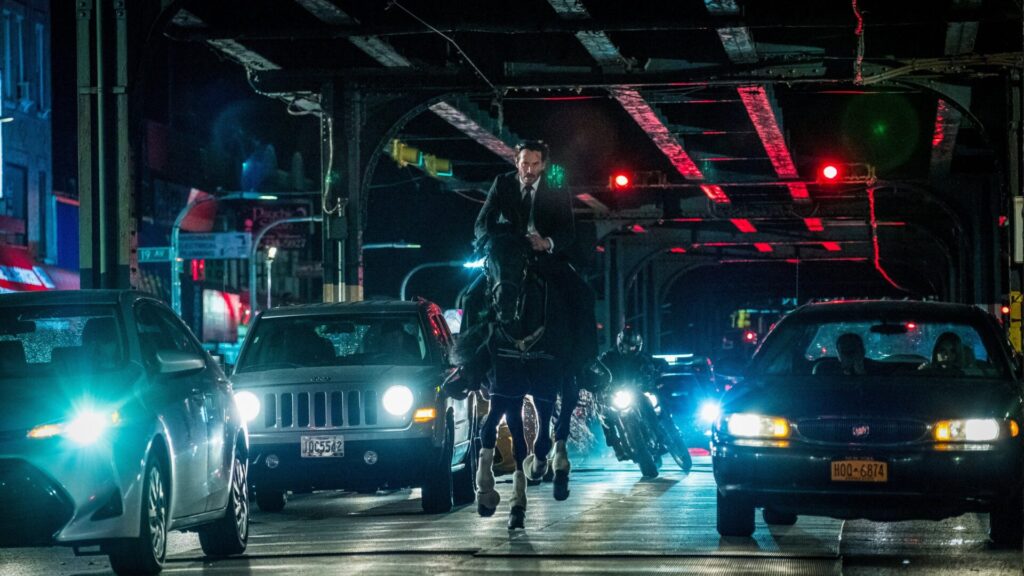 John Wick reitet auf einem Pferd in der Nacht in einer Stadt. Um ihn herum sind Autos und Motorräder zu sehen. Eine Top Fahrzeug-Action-Szene aus "John Wick. Kapitel 3".