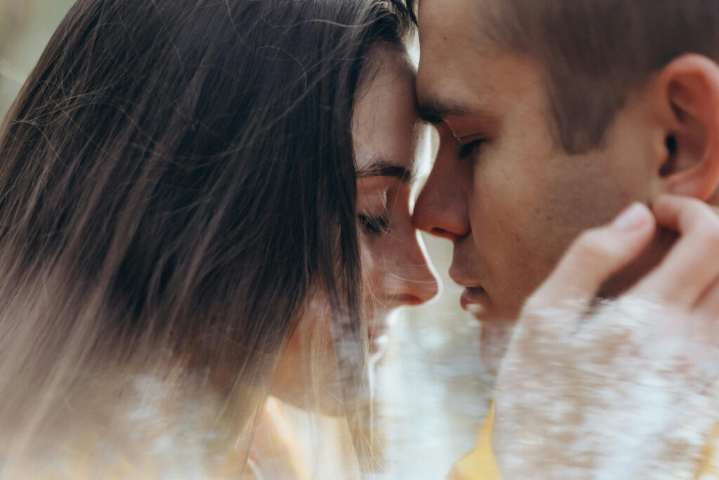 Nahaufnahme eines jungen Paares, das sich liebevoll gegenübersteht. Ihre Gesichter sind dicht beieinander, die Augen geschlossen, und sie berühren sich sanft. Die Szene vermittelt Intimität und emotionale Nähe. Ein weiches Licht und ein verschwommener Vordergrund erzeugen eine träumerische, romantische Atmosphäre.