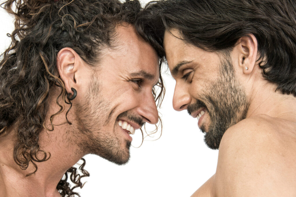 Zwei shirtlose Männer mit langen Haaren und Gesichtshaaren lächeln sich liebevoll an, während sie die Stirn aneinanderlegen. Vor weißem Hintergrund entsteht ein intimer Moment voller Zuneigung, Nähe und queerer Liebe. | Media Monday #739