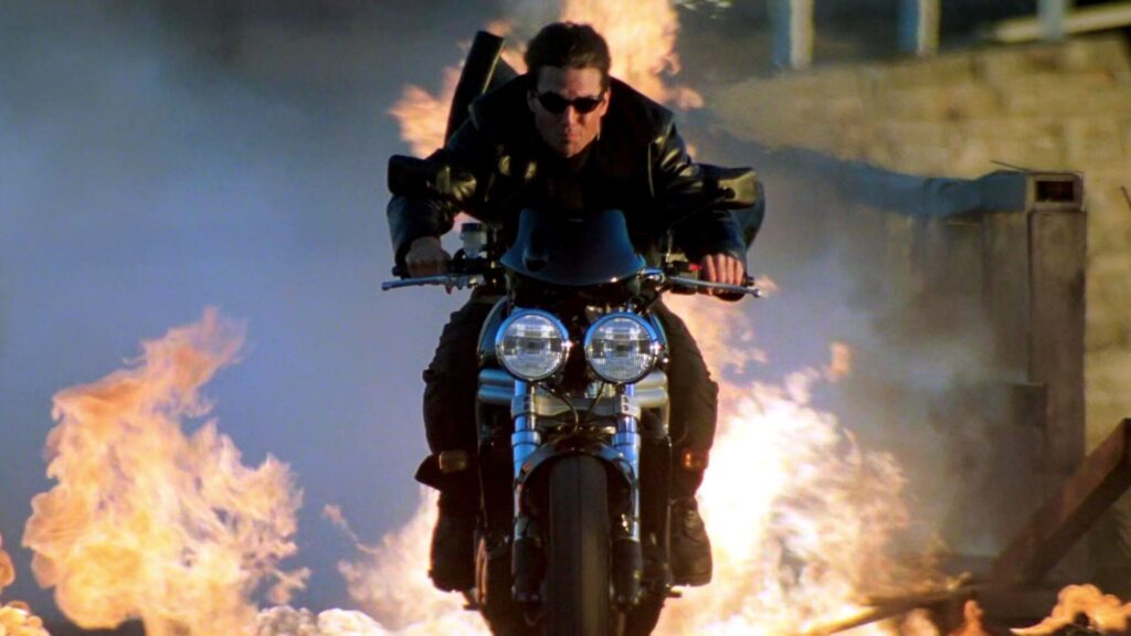 Tom Cruise sitz mit Sonnenbrille und Lederjacke auf einem Motorrad. Hinter ihm sind Flammen. Eine Top Fahrzeug-Action-Szene aus dem Film "Mission:Impossible II"