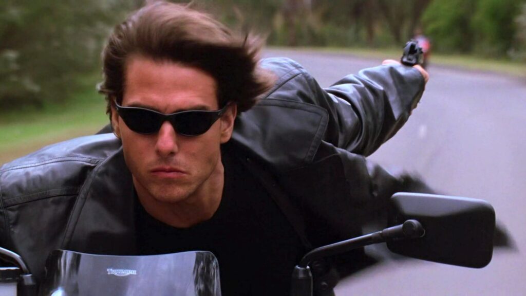 Tom Cruise fährt auf einem Motorrad. Ohne den Blick von der Straße abzuwenden, schießt er mit einer Pistole nach hinten. Top Fahrzeug-Action-Szene aus "Mission: Impossible II"
