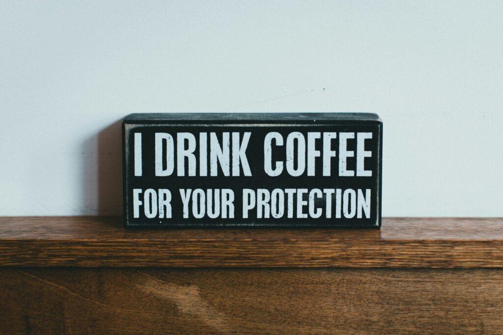 Schwarzes rechteckiges Schild mit weißer Aufschrift: ‚I DRINK COFFEE FOR YOUR PROTECTION‘ – auf einem Holztisch vor weißer Wand platziert.