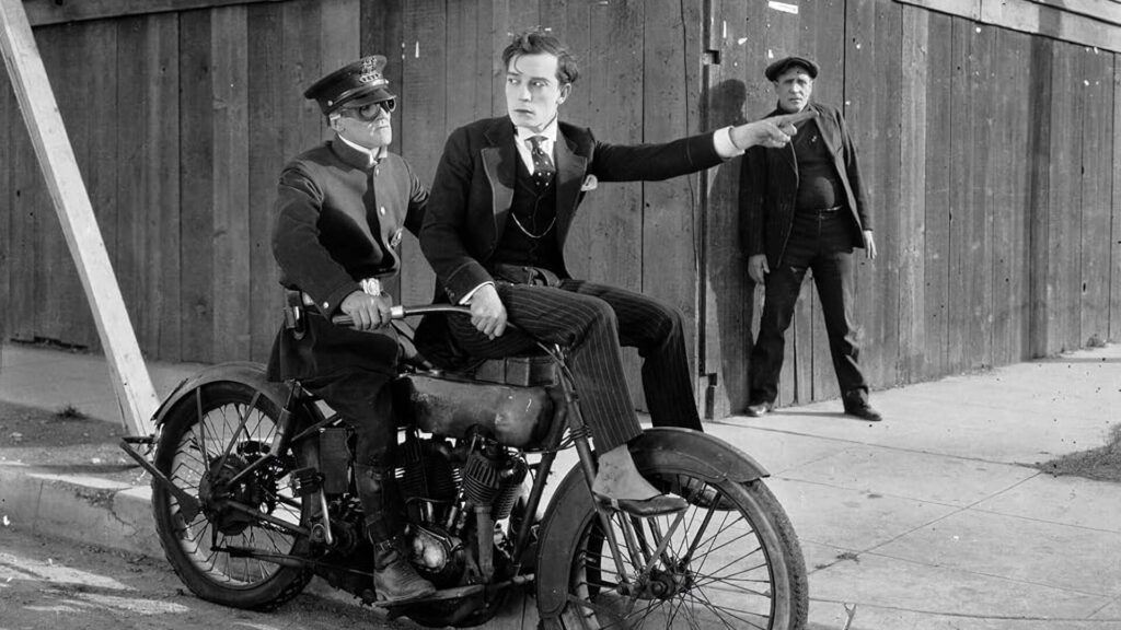 Buster Keaton sitzt auf dem Lenker eines Motorrades und weist den Weg an. Das Bild in schwarz-weiß stammt aus dem Film "Sherlock Jr.".