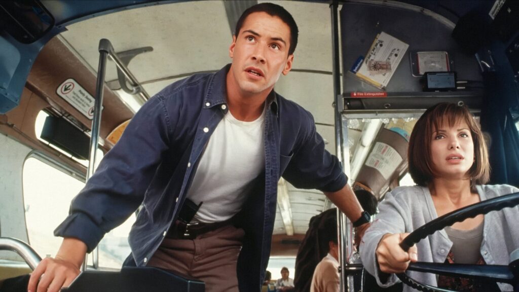 Keanu Reeves steht vorne im einem Bus. Neben ihm sitzt Sandra Bullock am Steuer. Top Fahrzeug-Action-Szene aus dem Film "Speed".