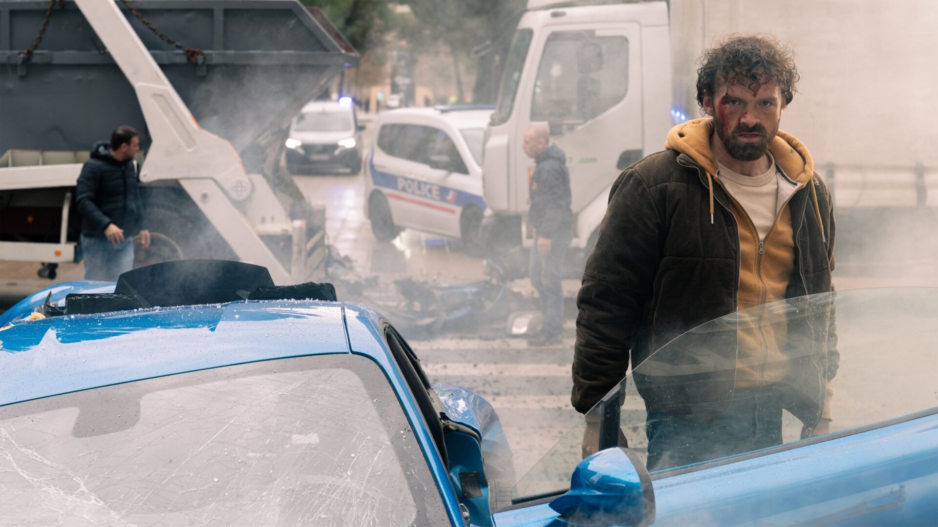 Nach einer Top Fahrzeug-Action-Szene steht Alban Lenoir neben einem zerstörten blauen Auto. Im Hintergrund sind Polizeiwagen und Rauch zu erkennen. Eine Top Fahrzeug-Action-Szene aus "Verirrte Kugel 3".